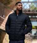 A man stands outside wearing the Regatta Essential Thermal Jacket by Regatta Professional, a black quilted coat made from recycled polyester, with his hands in the pockets and architectural elements softly blurred in natural light behind him.