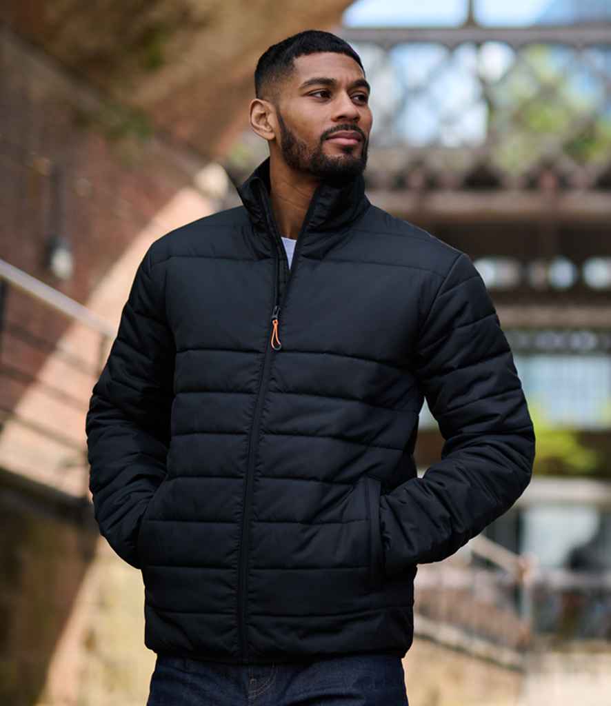 A man stands outside wearing the Regatta Essential Thermal Jacket by Regatta Professional, a black quilted coat made from recycled polyester, with his hands in the pockets and architectural elements softly blurred in natural light behind him.
