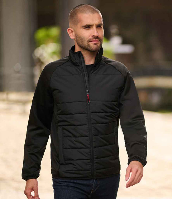 A man with a shaved head and trimmed beard walks outdoors in the Regatta Essential Hybrid Jacket by Regatta Professional, featuring quilted recycled polyester panels, paired with dark jeans. The blurred background shows stone structures and greenery.