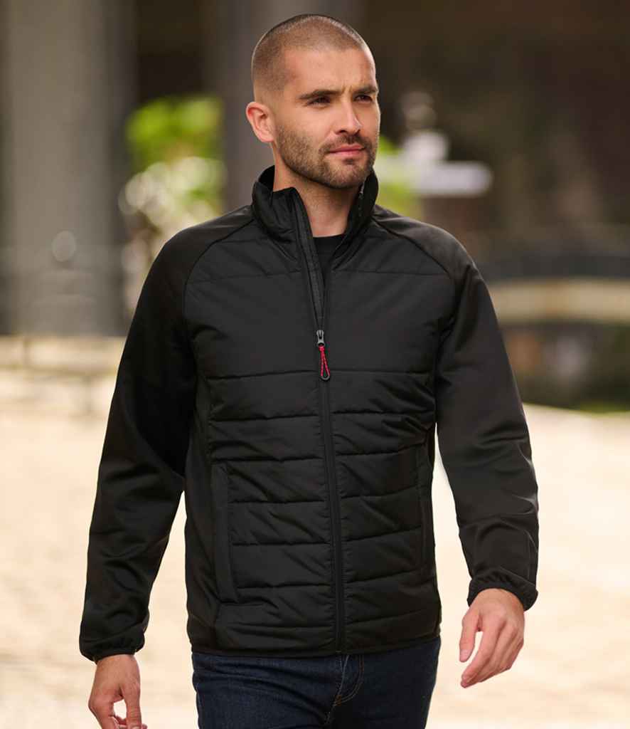 A man with a shaved head and trimmed beard walks outdoors in the Regatta Essential Hybrid Jacket by Regatta Professional, featuring quilted recycled polyester panels, paired with dark jeans. The blurred background shows stone structures and greenery.