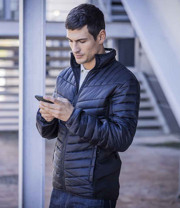 A man in a Regatta Professional Regatta Tourer Hybrid Jacket with Warmloft insulation stands outdoors, looking down at his smartphone against a backdrop of metal beams and horizontal slats.