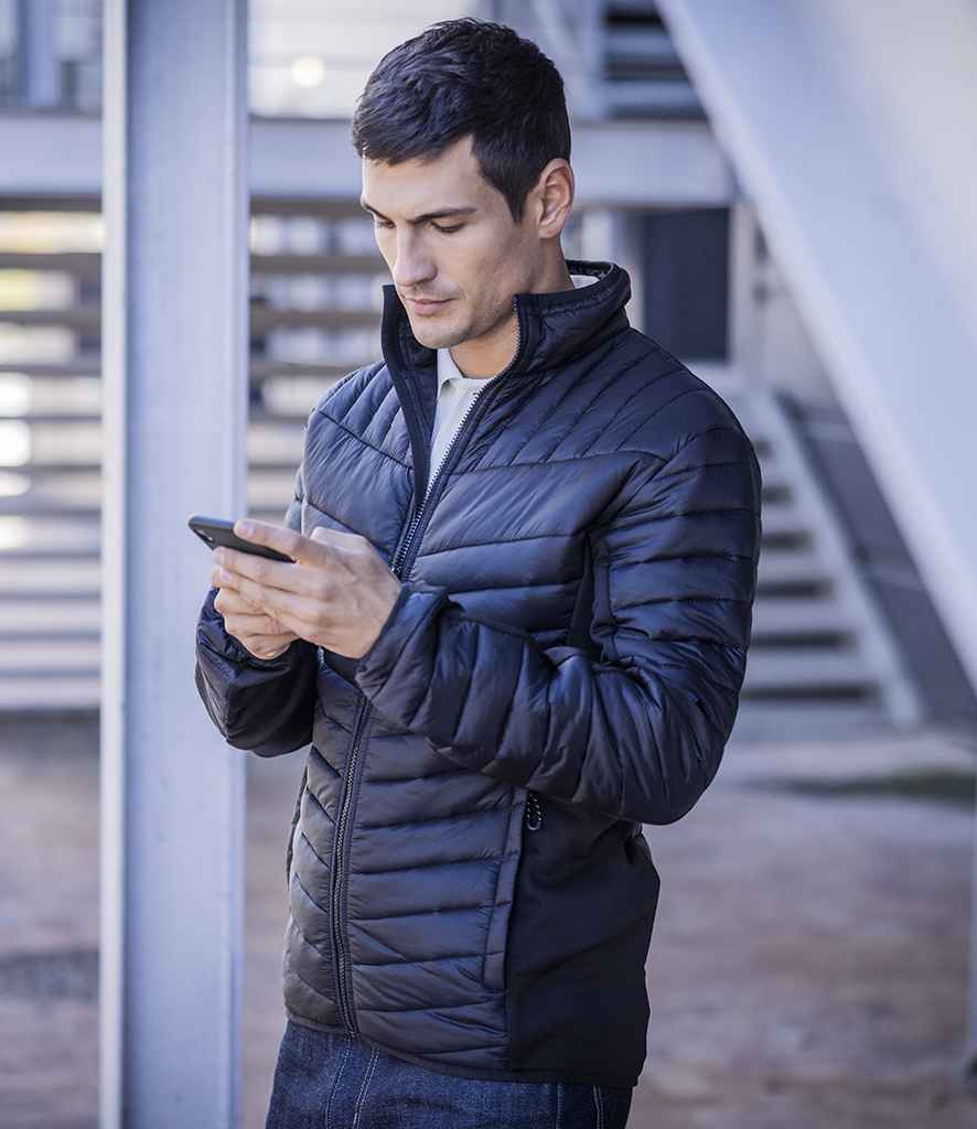 A man in a Regatta Professional Regatta Tourer Hybrid Jacket with Warmloft insulation stands outdoors, looking down at his smartphone against a backdrop of metal beams and horizontal slats.