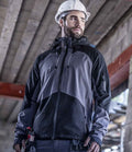 A man wearing a white construction helmet and the Regatta Professional 4TEX Stretch Waterproof Shell Jacket stands indoors at a construction site with tools in his pocket, glancing sideways. Unfinished concrete beams are visible in the background.