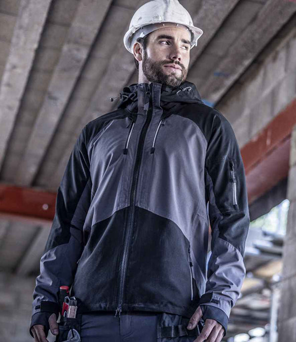A man wearing a white construction helmet and the Regatta Professional 4TEX Stretch Waterproof Shell Jacket stands indoors at a construction site with tools in his pocket, glancing sideways. Unfinished concrete beams are visible in the background.