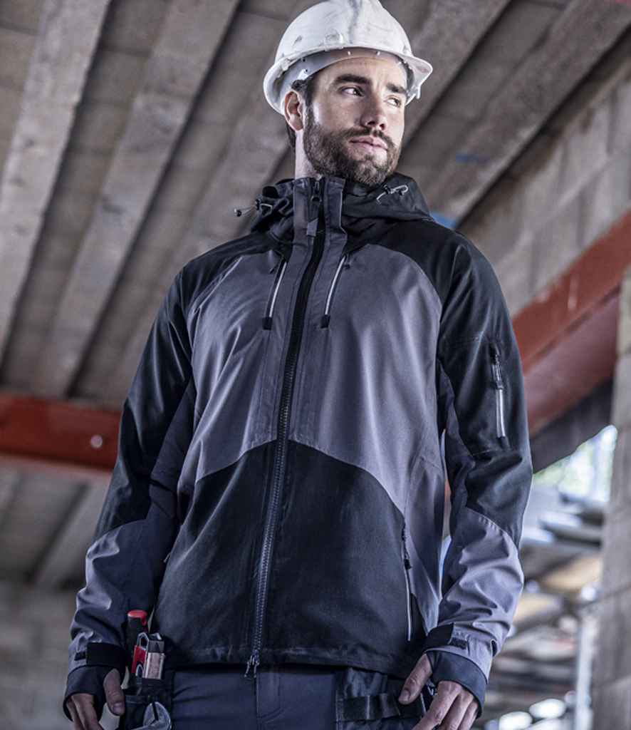 A man wearing a white construction helmet and the Regatta Professional 4TEX Stretch Waterproof Shell Jacket stands indoors at a construction site with tools in his pocket, glancing sideways. Unfinished concrete beams are visible in the background.