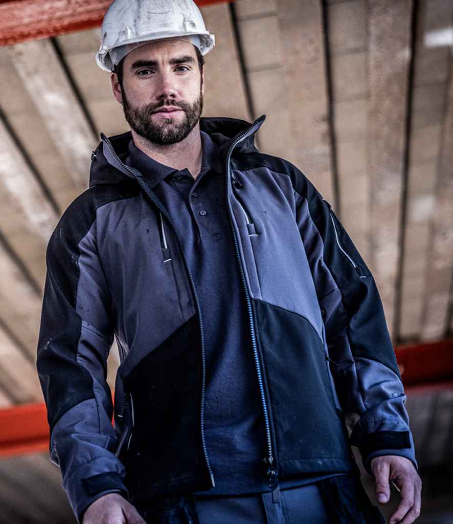 A man wearing a white construction helmet and the Regatta Professional Regatta 4TEX Stretch Three Layer Soft Shell Jacket stands indoors under a wooden ceiling, looking slightly down at the camera.