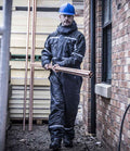 A person in a blue hard hat and Regatta Professional Regatta Pro Waterproof Insulated Coverall walks outdoors between a brick wall and fence, carrying several copper pipes.