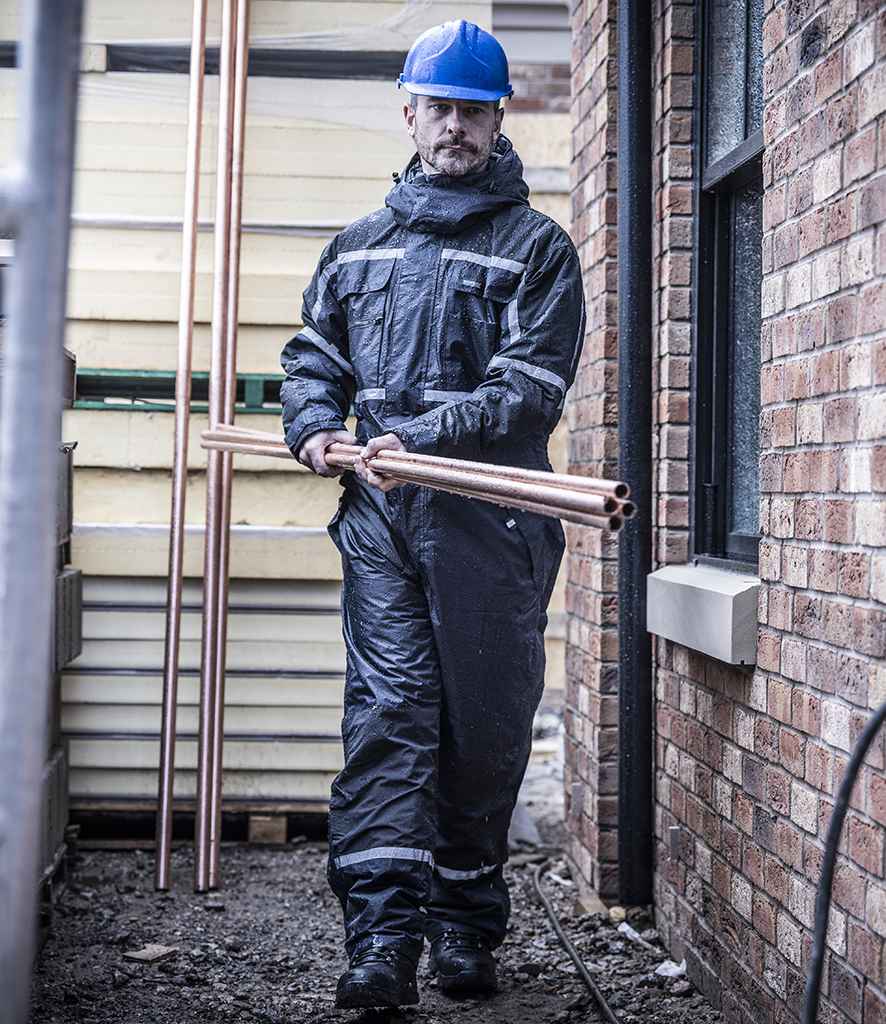 A person in a blue hard hat and Regatta Professional Regatta Pro Waterproof Insulated Coverall walks outdoors between a brick wall and fence, carrying several copper pipes.