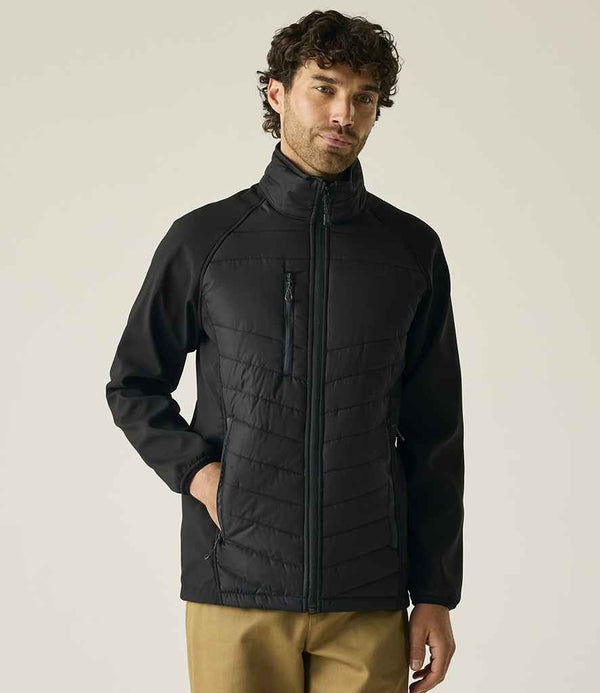 A man stands against a plain background wearing the Regatta Pro Universal Jacket from Regatta Professional, a black zip-up stretch soft shell with a high collar, paired with khaki pants. His left hand is in his pocket and his right hand is by his side.