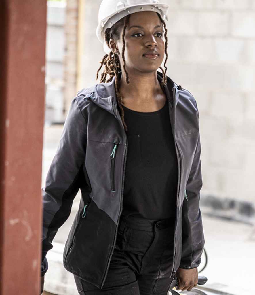 A person wearing a white hard hat, black shirt, and a Regatta Professional Ladies Ada Stretch Soft Shell Jacket stands indoors near a partially constructed wall.