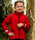 A young child with brown hair tied back stands outdoors by a tree, wearing the Result Kids Classic Soft Shell Jacket by Result. The bright red jacket contrasts with the lush greenery in the background as the child looks at the camera.