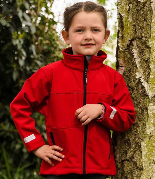 A young child with brown hair tied back stands outdoors by a tree, wearing the Result Kids Classic Soft Shell Jacket by Result. The bright red jacket contrasts with the lush greenery in the background as the child looks at the camera.