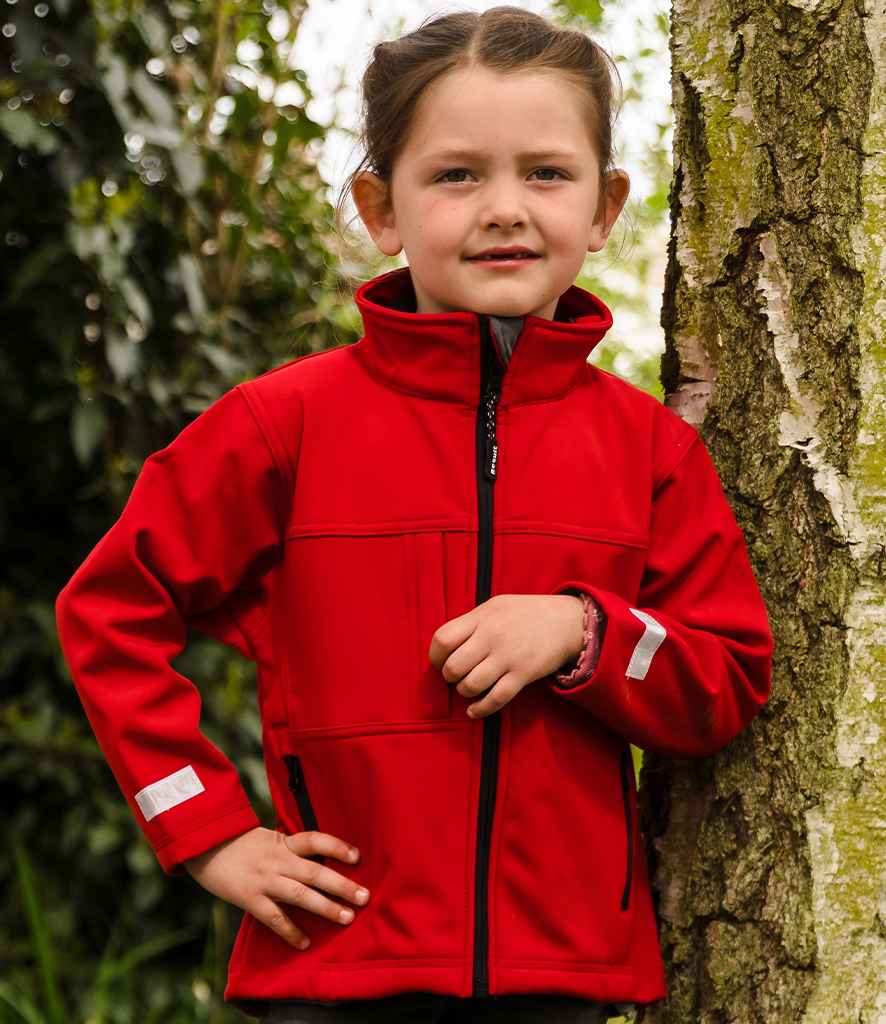 A young child with brown hair tied back stands outdoors by a tree, wearing the Result Kids Classic Soft Shell Jacket by Result. The bright red jacket contrasts with the lush greenery in the background as the child looks at the camera.