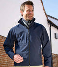 A man wearing the Result Classic Soft Shell Jacket by Result and a striped scarf stands outside in front of a white building with a brick lower half, looking to the side and smiling.