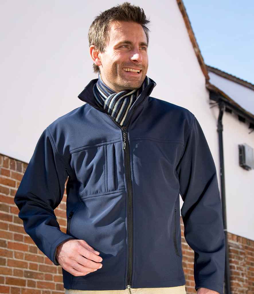 A man wearing the Result Classic Soft Shell Jacket by Result and a striped scarf stands outside in front of a white building with a brick lower half, looking to the side and smiling.
