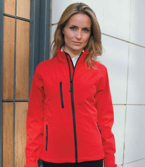 A woman with light brown hair stands before a tiled wall, wearing the Result Ladies Base Layer Soft Shell Jacket—a bright red, lightweight, showerproof zip-up by Result with black zippers—over a white shirt. She faces the camera neutrally.