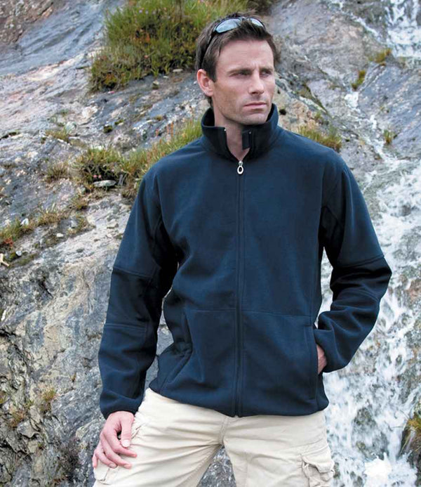 A man stands outdoors before rocks and a small waterfall, sunglasses on his head, wearing the Result Osaka Combed Pile Soft Shell Jacket by Result with light-colored cargo shorts.