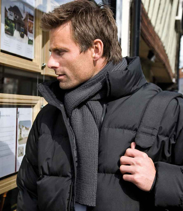 A man wearing a black, tag free puffer jacket and a dark Result Classic Heavy Knit Scarf from Result Winter Essentials stands outside, looking at real estate listings in a window with a black backpack over one shoulder.