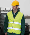 A man wearing a yellow hard hat and a Result Core Motorist Hi-Vis Vest with reflective stripes stands outdoors before a dark green off-road vehicle, with water, land, and an overcast sky in the background.