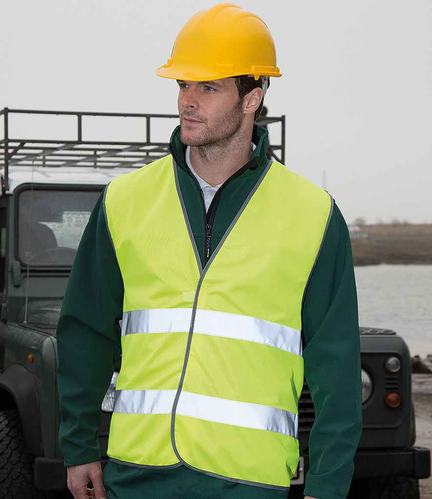A man wearing a yellow hard hat and a Result Core Motorist Hi-Vis Vest with reflective stripes stands outdoors before a dark green off-road vehicle, with water, land, and an overcast sky in the background.
