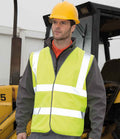 A man in a yellow safety helmet and a Result Core Hi-Vis Motorway Vest by Result Core stands before construction equipment, wearing a gray jacket, at a construction site.