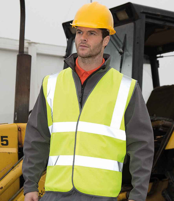 A man in a yellow safety helmet and a Result Core Hi-Vis Motorway Vest by Result Core stands before construction equipment, wearing a gray jacket, at a construction site.