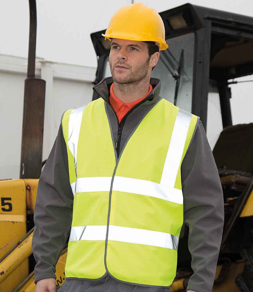 A man in a yellow safety helmet and a Result Core Hi-Vis Motorway Vest by Result Core stands before construction equipment, wearing a gray jacket, at a construction site.