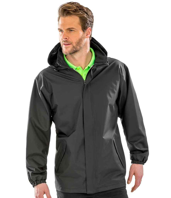 A man wearing the Result Core Midweight Jacket by Result Core, featuring a black color and adjustable hood, stands facing slightly to the side over a bright green collared shirt with his hands at his sides.