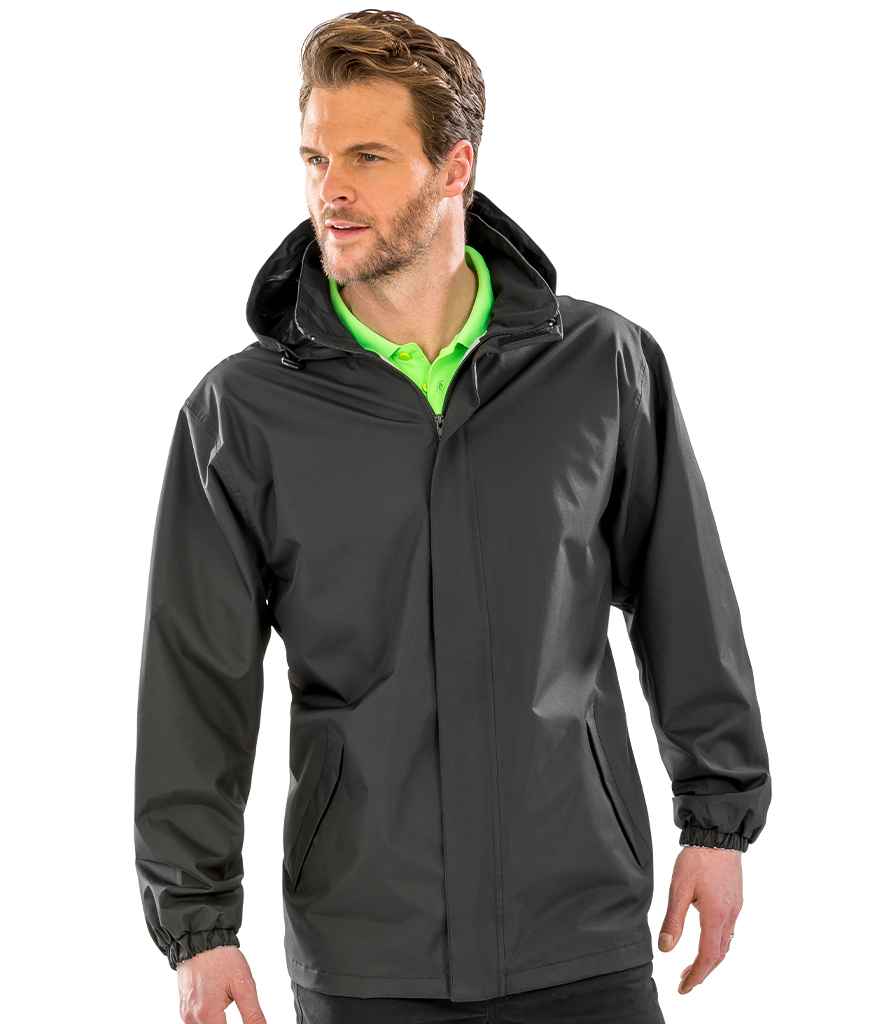 A man wearing the Result Core Midweight Jacket by Result Core, featuring a black color and adjustable hood, stands facing slightly to the side over a bright green collared shirt with his hands at his sides.