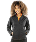 A person with curly hair is smiling and standing with their hands in the pockets of a black Result Core Ladies Soft Shell Jacket, worn over an orange collared shirt. They are looking slightly to the side.