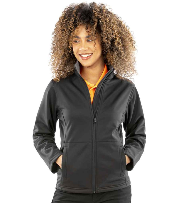 A person with curly hair is smiling and standing with their hands in the pockets of a black Result Core Ladies Soft Shell Jacket, worn over an orange collared shirt. They are looking slightly to the side.
