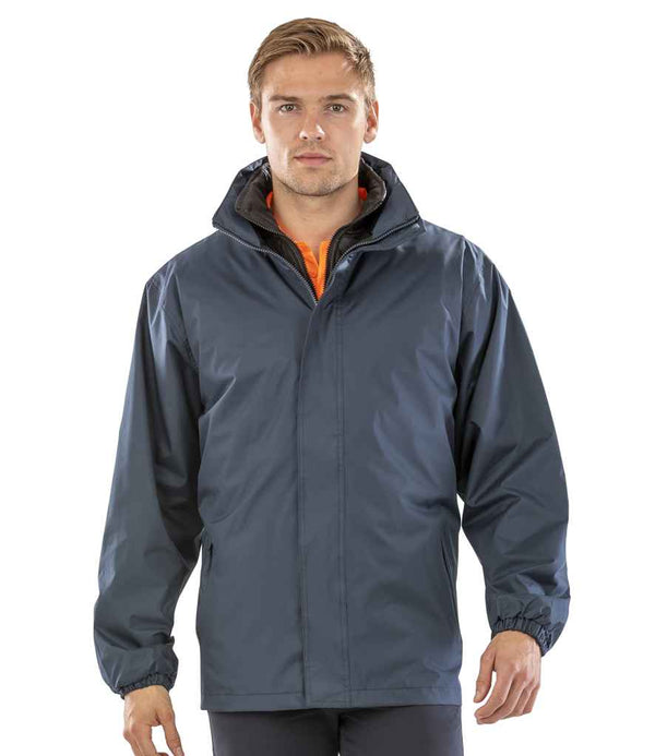 A person stands facing the camera in a Result Core 3-in-1 Jacket by Result Core, dark blue with a hood, zipper, and elastic cuffs, worn over an orange shirt. The background is plain white.