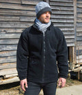 A man wearing a gray knit beanie, matching scarf, black pants, and a Result Core Polartherm™ Quilted Winter Fleece Jacket from Result Core stands outside in front of a weathered wooden wall.