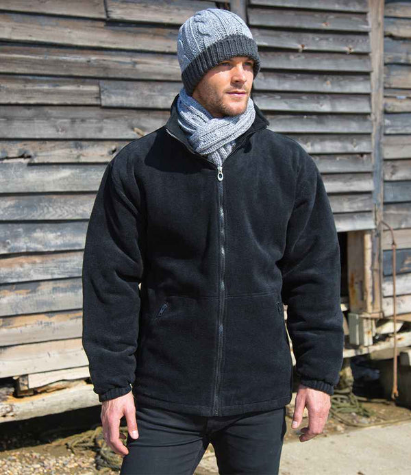 A man wearing a gray knit beanie, matching scarf, black pants, and a Result Core Polartherm™ Quilted Winter Fleece Jacket from Result Core stands outside in front of a weathered wooden wall.