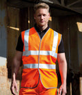 A man with short, light hair stands outdoors before a weathered metal structure, wearing a bright orange Result Safe-Guard Hi-Vis Vest with reflective stripes from Result Safe-Guard and looking directly at the camera.