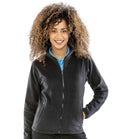 A woman with curly hair stands smiling, wearing the Result Core Ladies Norse Outdoor Fleece—a black fitted jacket—over a blue collared shirt, with one hand in her pocket, against a plain white background.