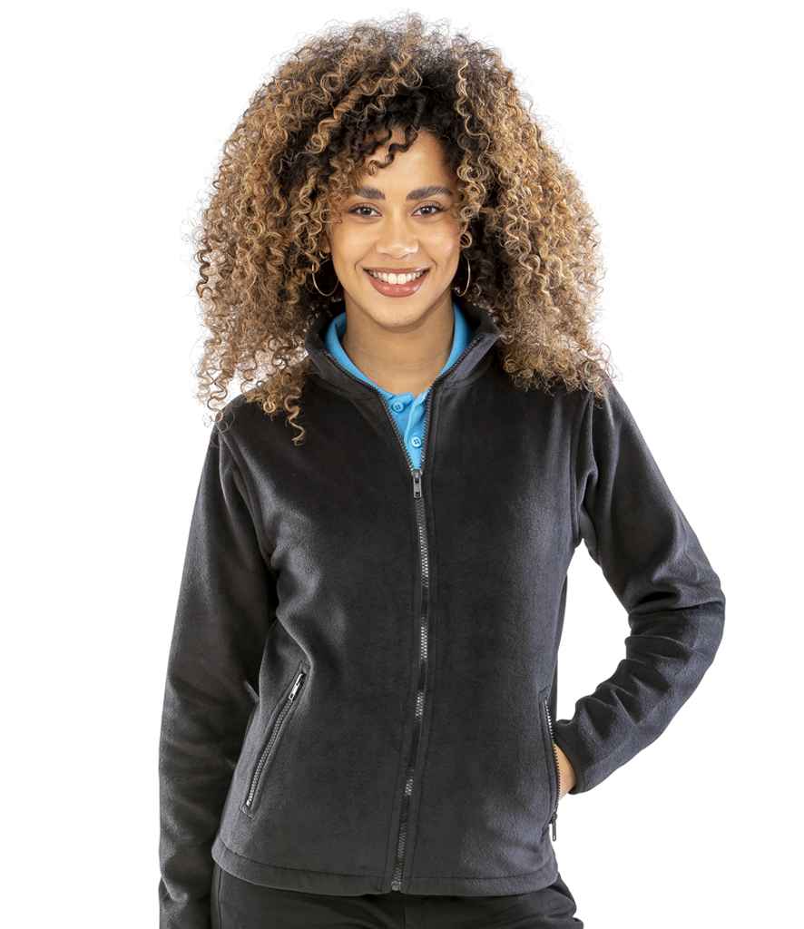 A woman with curly hair stands smiling, wearing the Result Core Ladies Norse Outdoor Fleece—a black fitted jacket—over a blue collared shirt, with one hand in her pocket, against a plain white background.