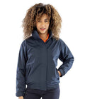 A person with curly hair stands facing forward, wearing a Result Core Ladies Channel Jacket by Result Core over an orange shirt, with their left hand on their hip.