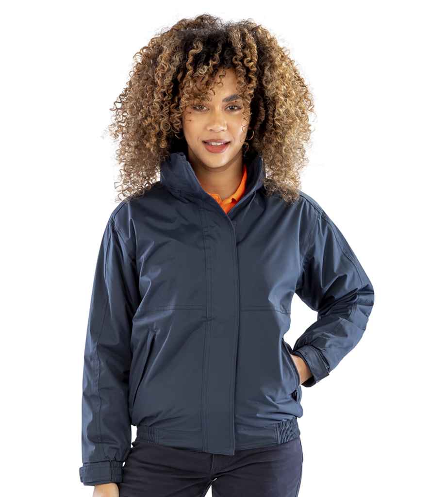 A person with curly hair stands facing forward, wearing a Result Core Ladies Channel Jacket by Result Core over an orange shirt, with their left hand on their hip.