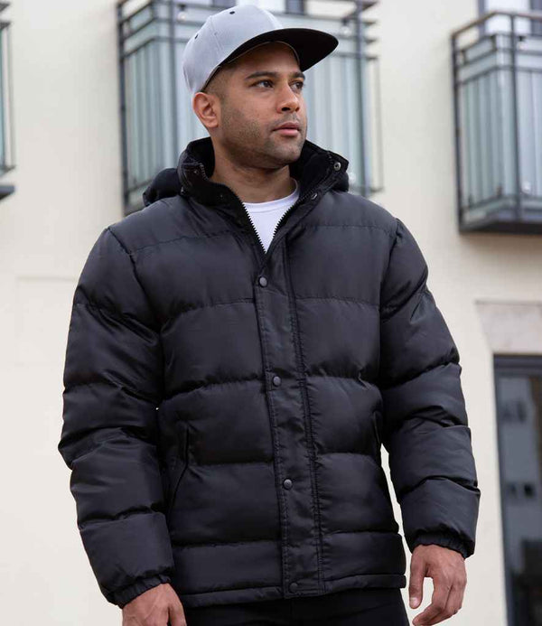A man wearing a Result Core Nova Lux Padded Jacket and a gray baseball cap stands outside in front of a building with balconies, looking to the side.