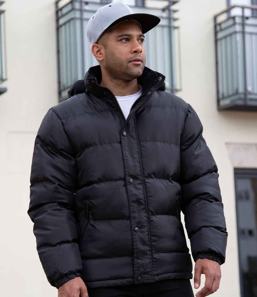 A man wearing a Result Core Nova Lux Padded Jacket and a gray baseball cap stands outside in front of a building with balconies, looking to the side.