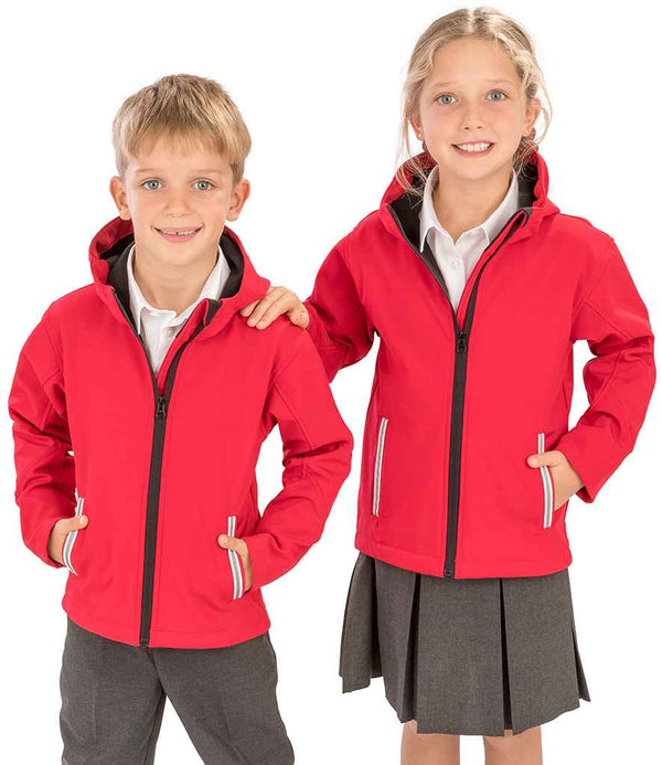 Two children smile at the camera wearing Result Core Kids TX Performance Hooded Soft Shell Jackets by Result Core over white collared shirts; she pairs hers with a grey skirt, he wears grey shorts. Both have hands tucked into their jacket pockets.
