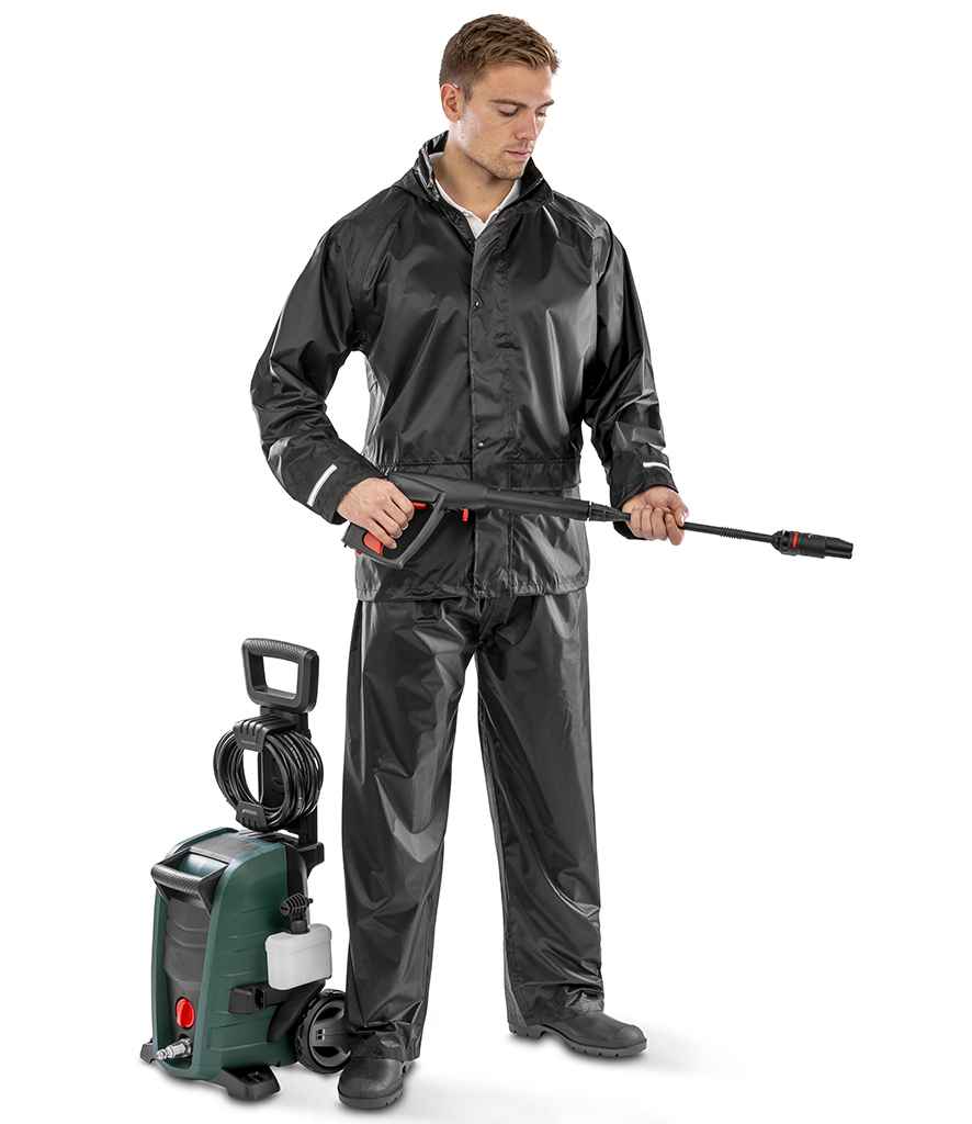 A man wearing a Result Core Rain Suit by Result Core with reflective trim and black shoes stands holding a pressure washer wand. A green pressure washer with a hose and wheels is next to him on the floor.