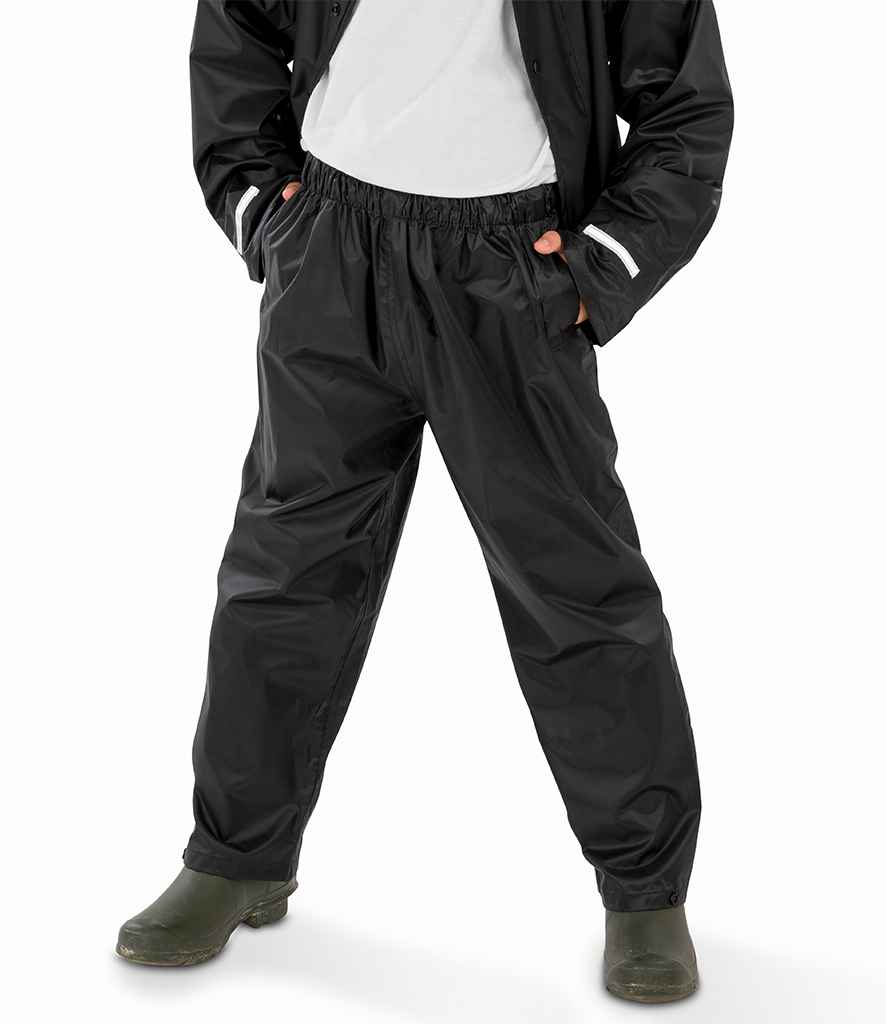 A person wearing Result Core Kids Waterproof Overtrousers by Result Core with an elasticated waistband, a black jacket, a white shirt, and green rubber boots stands with hands in pockets against a white background.
