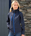 A person with long blonde hair wears the Result Core Ladies Hooded Soft Shell Jacket by Result Core, featuring a navy blue waterproof design, high collar, blue zippers, and fleece lining, standing before a stone wall and dark blue door.
