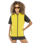 A person with curly hair wears brown sunglasses and the Result Core Ladies Printable Soft Shell Bodywarmer over a short-sleeved black top, smiling with one hand on their hip against a plain white background.
