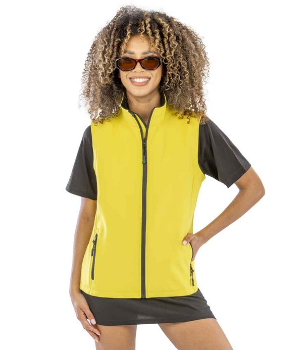 A person with curly hair wears brown sunglasses and the Result Core Ladies Printable Soft Shell Bodywarmer over a short-sleeved black top, smiling with one hand on their hip against a plain white background.