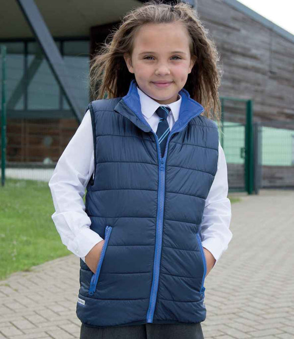 A young girl smiles outdoors in front of a modern building, hands in her Result Core Kids Padded Bodywarmer pockets, layered over a white shirt, navy tie, and gray skirt.