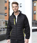 A man with short hair and a trimmed beard stands outside, wearing the Result Recycled Compass Padded Jacket by Result Recycled—black with yellow zipper accents, made with recycled padding—over a white shirt. Apartment buildings and a railing are visible behind him.