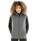 A person with curly hair stands against a white background, smiling at the camera in a gray Result Recycled Promo Padded Bodywarmer over a black long-sleeve top, hands tucked in the vest pockets.